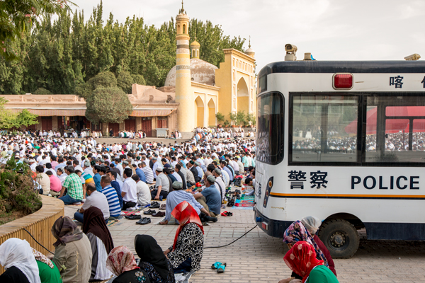 Temmuz 2015'te, antik İpek Yolu ticaret kasabası Kaşgar'daki Id Kah Camii'nin dışında, Uygur kadınları ve erkekler Ramazan'ın sonunu kutlayan neşeli bir Müslüman bayramı sırasında dua ediyorlar.  Polis araçları ve güvenlik hattı Xinjiang, Çin'deki kamu meydanında.  Alexandra Williams