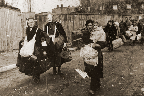 Elderly women carrying young children and bundles of personal belongings trudge along a street in the Łódź ghetto toward the assembly point for deportations to Chełmno. US Holocaust Memorial Museum, courtesy of Muzeum Sztuki w Łodzi