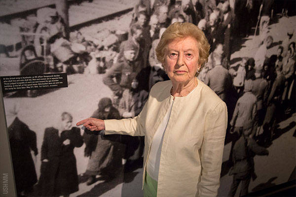 Irene Weiss, a Holocaust survivor and Museum volunteer, stands in front of a photograph of herself when she was a child arriving at Auschwitz-Birkenau, on display in the Museum&rsquo;s main exhibition. USHMM (historic image Yad Vashem)